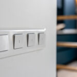 Close view of four switches on a white wall, crisp edges and matte plastic, a ladder and kitchen blur in the background, a simple daily tech moment.